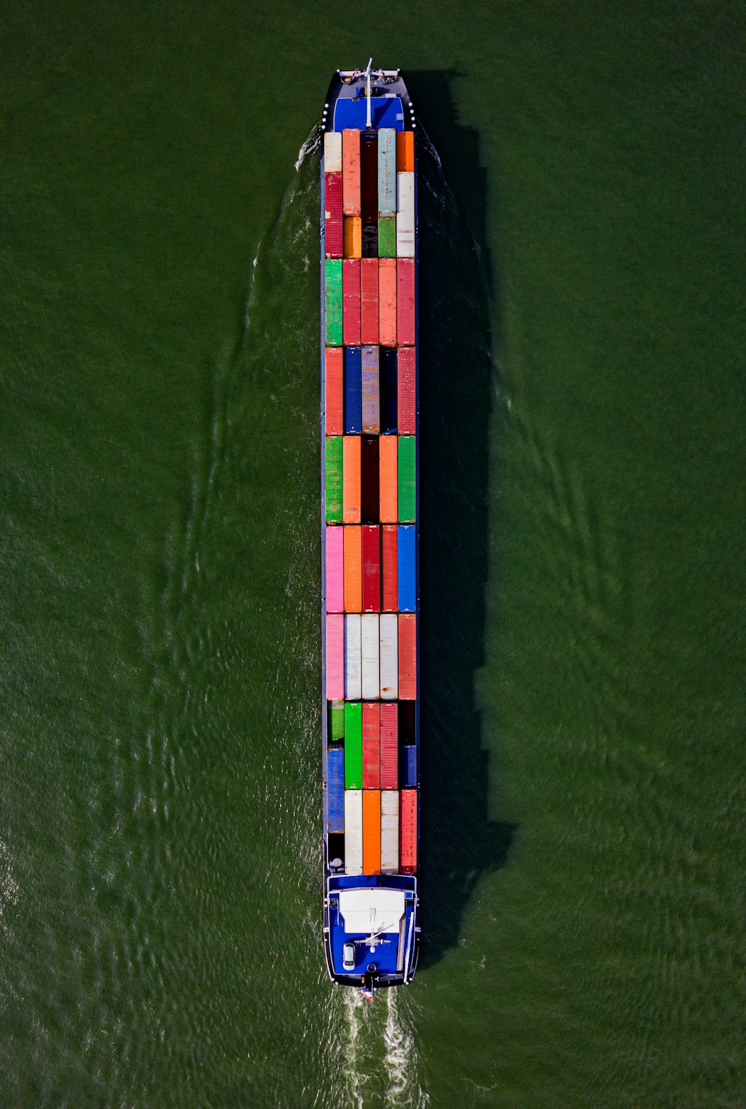 Captured from above, a cargo barge adorned with a mosaic of colorful containers makes its way down a river, vivid against the waters surface, exemplifying the essential role of inland waterways in global trade and logistics.